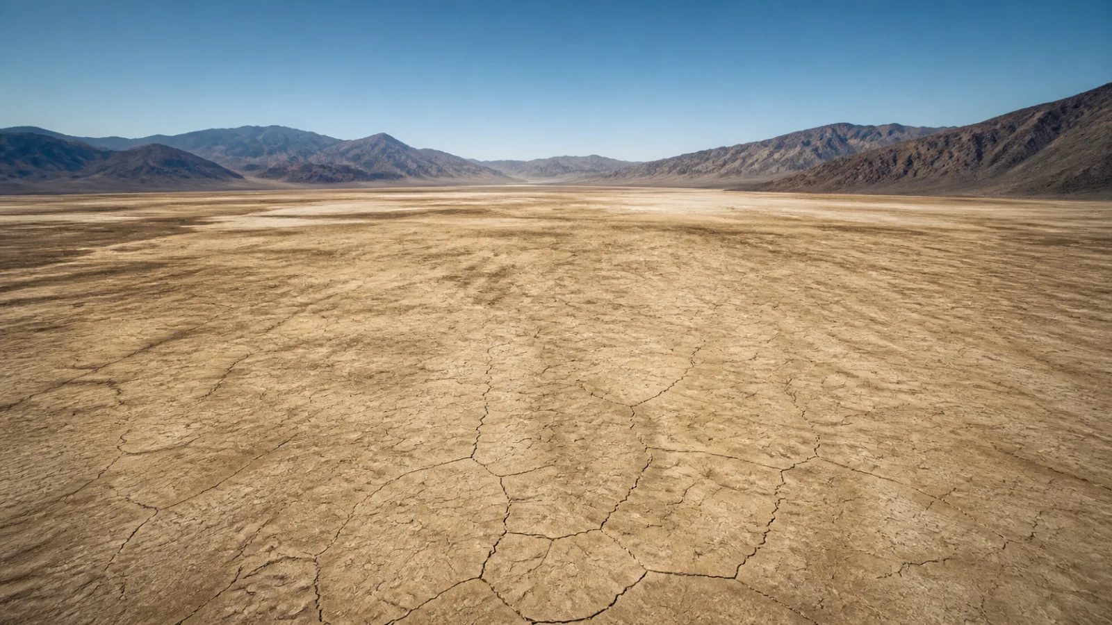 Wide aerial view of a vast pale cracked dry lakebed stretching toward distant brown desert mountains under a harsh sun